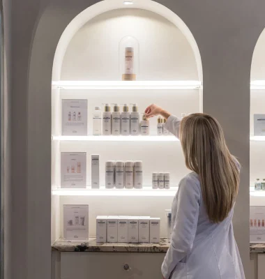 Woman in white coat examining skincare products on illuminated shelves in medical spa or clinic