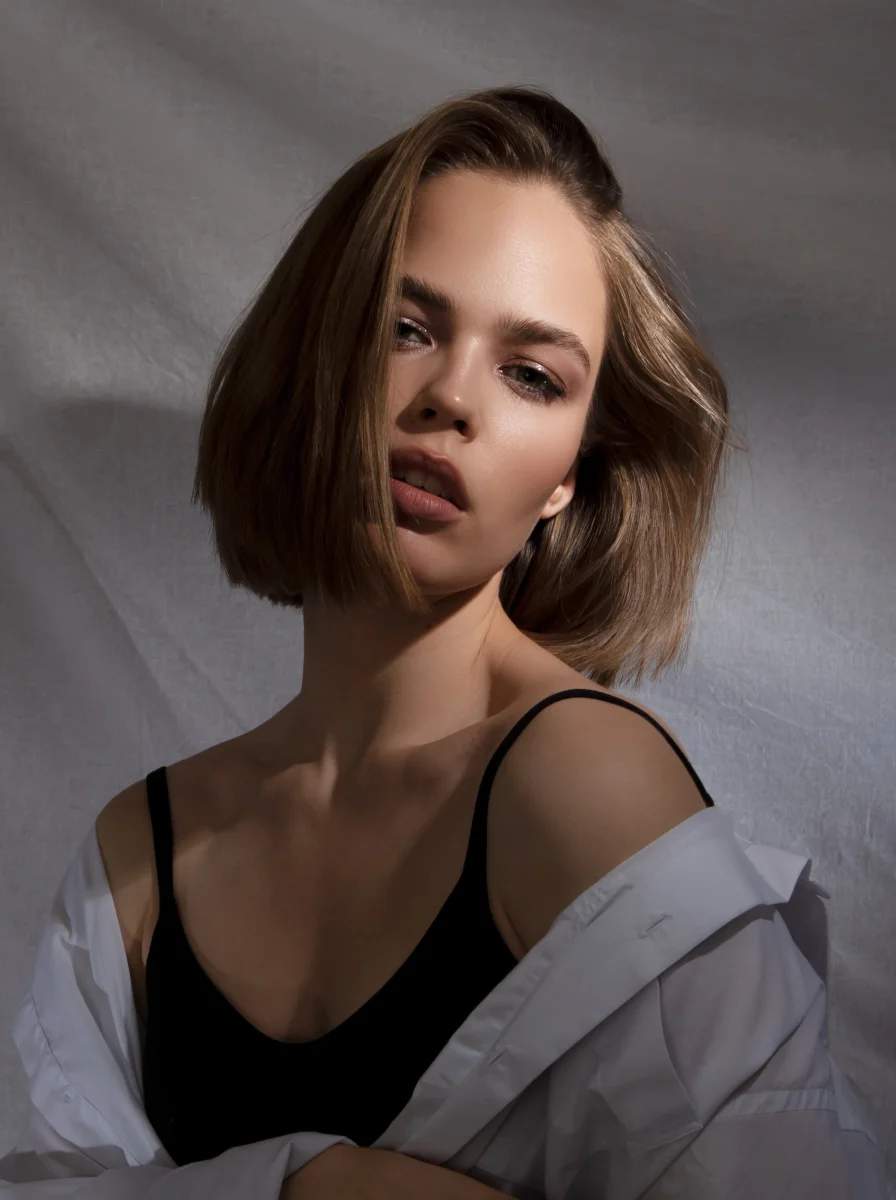 Young woman with shoulder-length hair in black tank top and white shirt against gray wall with dramatic lighting
