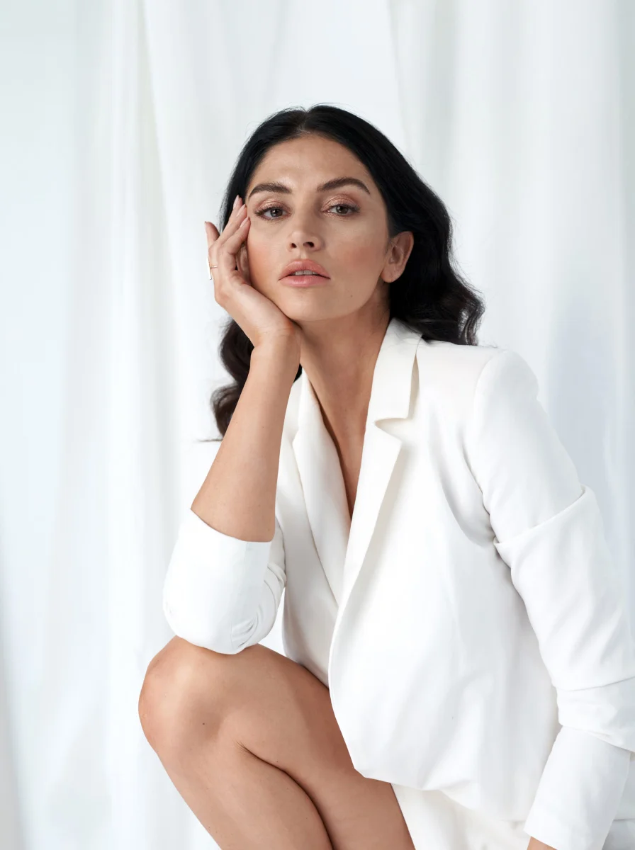 Woman in elegant white blazer posing gracefully against white curtain background in modern minimalist setting