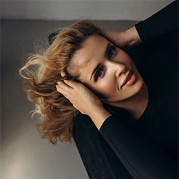 Woman with voluminous blonde curly hair in black turtleneck posing with hands near face against gray background