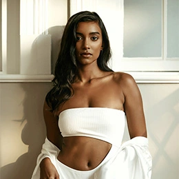 Woman in white bandeau top and pants posing against modern interior wall with natural lighting