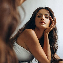 Woman with long dark hair in white dress looking at mirror reflection while touching her face