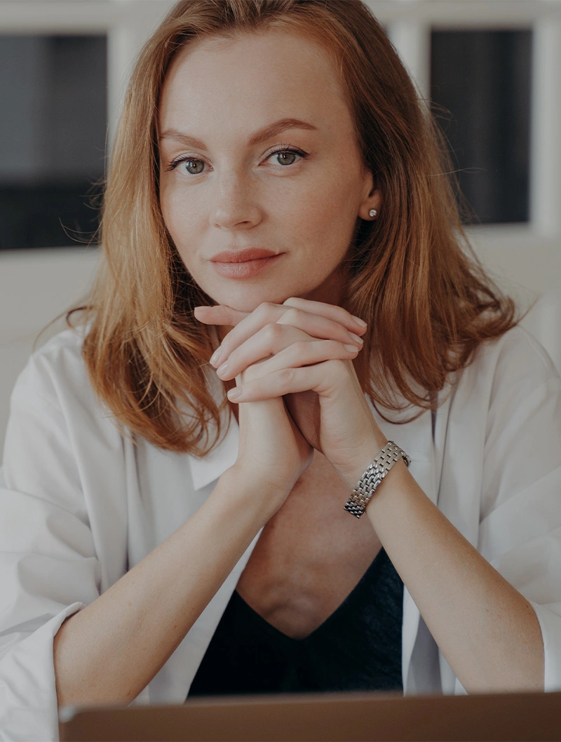 Professional woman with red hair in white blazer posing with hands clasped under chin in business portrait