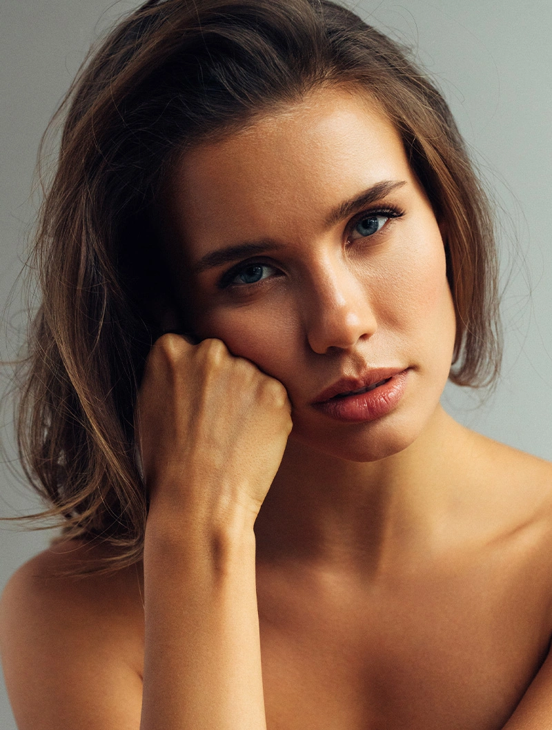 Woman with brown hair resting chin on hand, looking directly at camera with natural makeup and bare shoulders