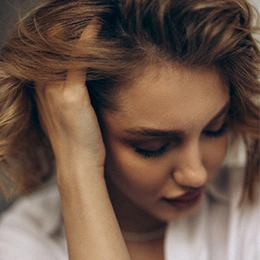Woman with tousled blonde hair looking down pensively with hand near face in soft natural lighting