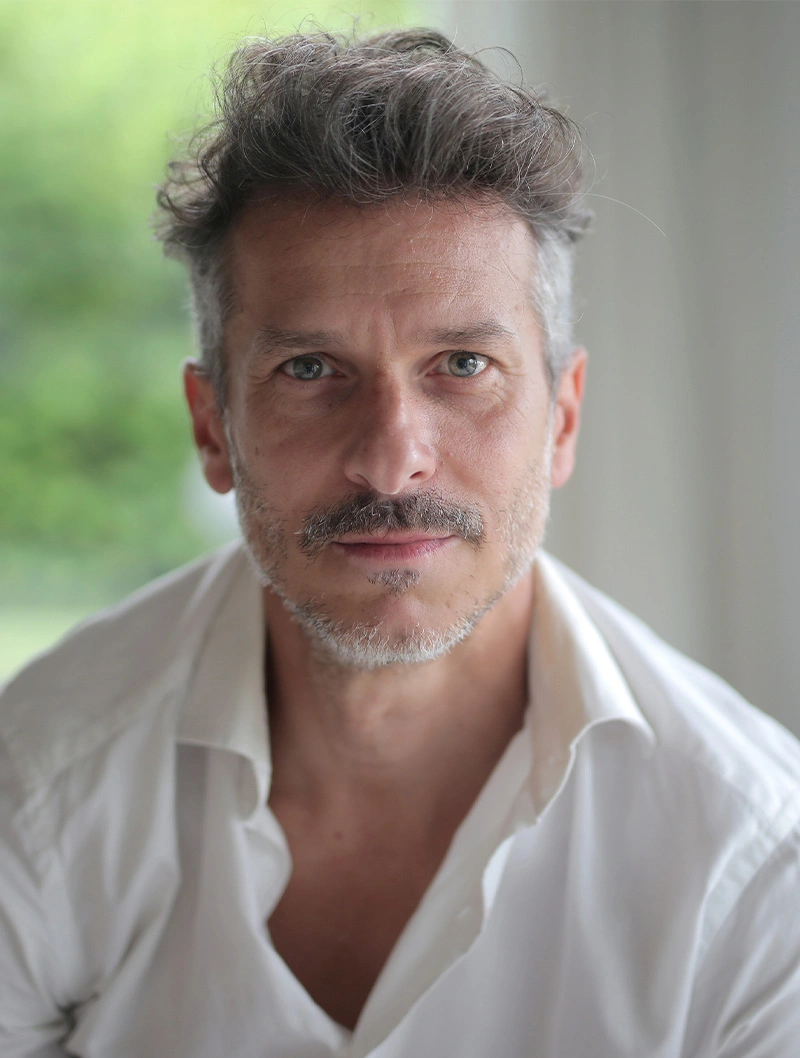 Professional headshot of mature man with gray hair and mustache wearing white shirt against blurred background