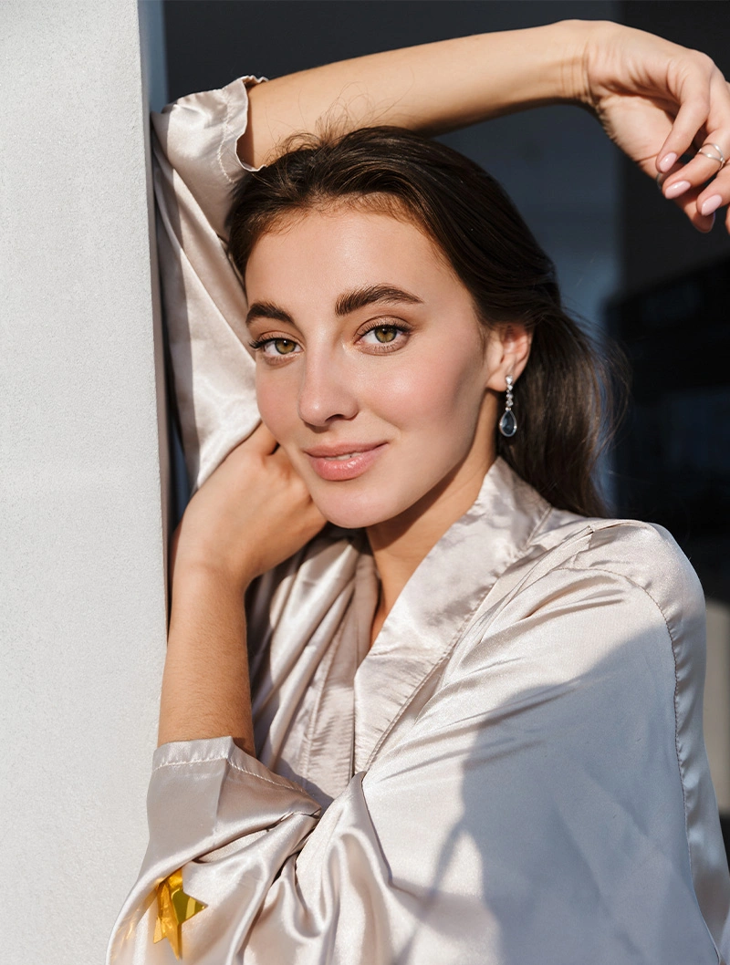 Woman in white blazer posing elegantly with hand near face against neutral background in natural lighting