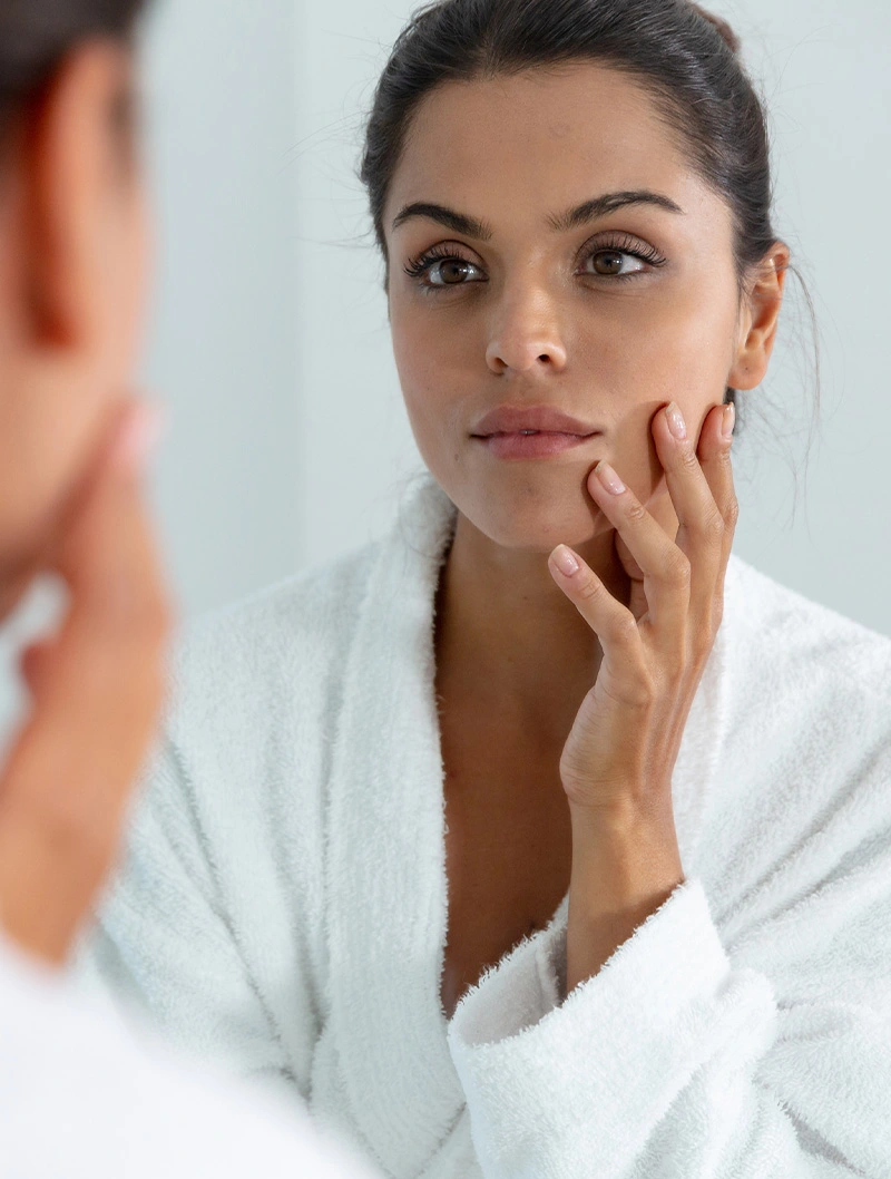 Woman examining her facial skin in mirror while touching face in white bathrobe