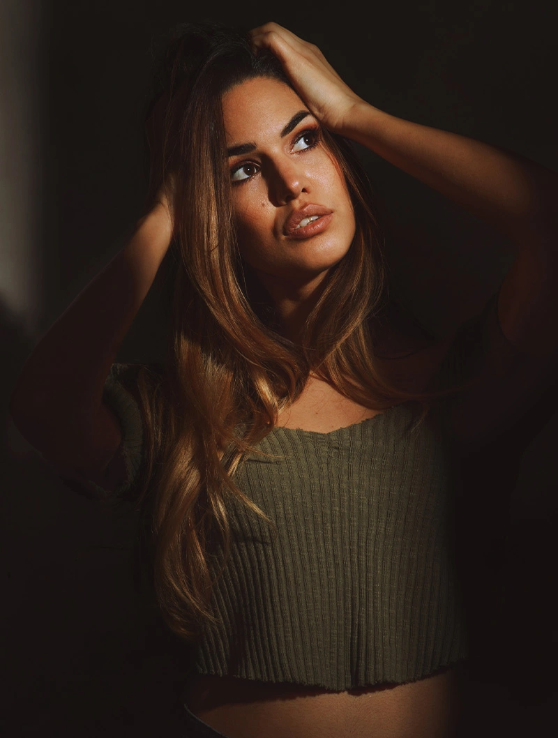 Young woman with long brown hair in olive green sweater posing with hands in hair against dark background