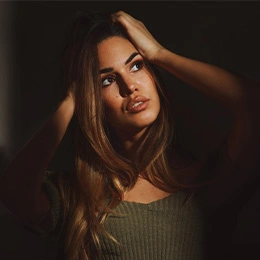 Woman with long brown hair in green sweater posing with hand in hair against dark background