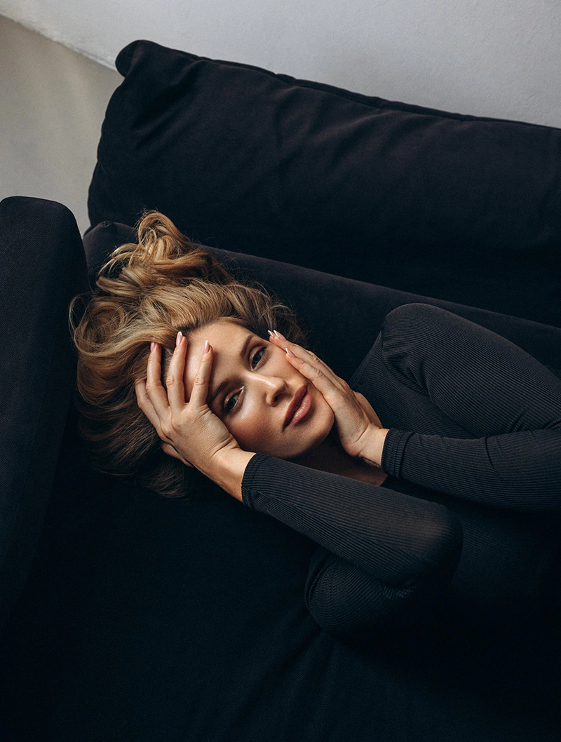 Young man with tousled blonde hair lying on black couch in dark clothing, hand on face in contemplative pose