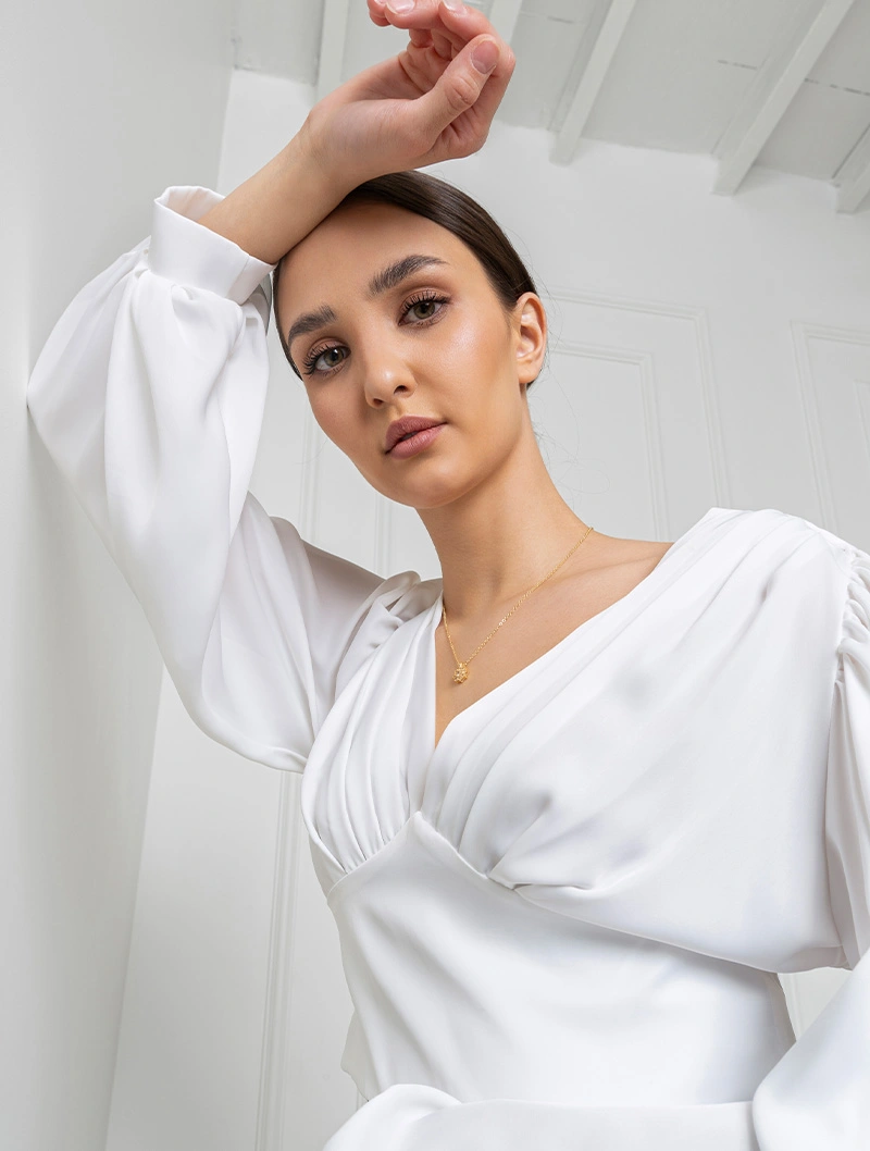 Woman in elegant white puff sleeve blouse posing with hand raised in minimalist white interior setting
