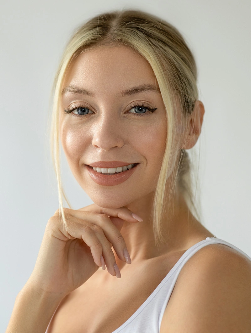 Smiling young woman with blonde hair in white tank top touching chin on neutral background
