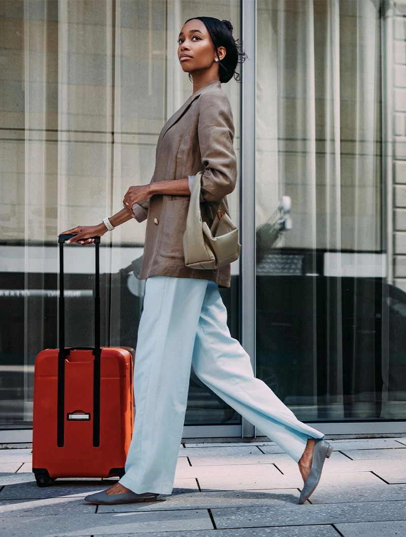 Woman in beige blazer pulling red suitcase while walking on urban street, dressed in casual travel outfit